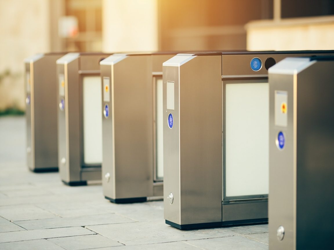 A row of stainless steel automated border gates with fingerprint pads and frosted glass panels, in soft morning light.